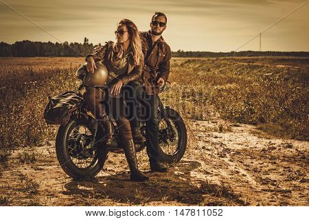 Young, stylish cafe racer couple on the vintage custom motorcycles in a field.
