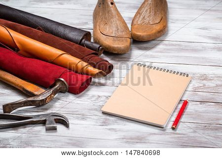 Brightly colored leather in rolls working tools shoe lasts notebook with pencil on white wooden background. Leather craft.