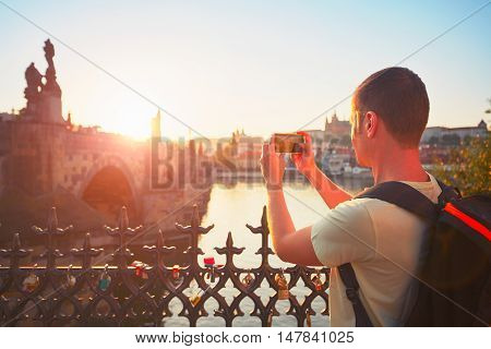 Traveler with backpack taking photo by mobile phone. Young tourist near Charles bridge at the sunset. Prague Czech Republic. - back lit