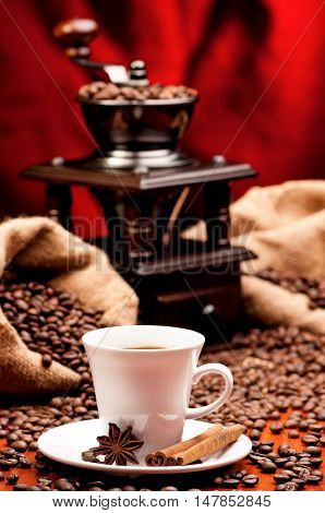 Cup of warm coffee with coffee grinder and burlap sack with roasted beans. Old coffee mill with white mug and spice - cinnamon and anise.