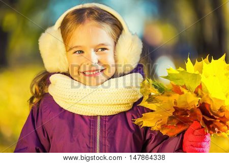Happy little girl in earflaps with autumn leaves in the park