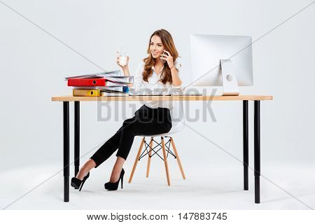 Beautiful businesswoman sitting at the desk with water glass in hand and talking on the phone isoltaed on the white background