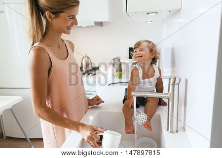 Mom with her 1,4 years old child washing dishes in the white kitchen interior