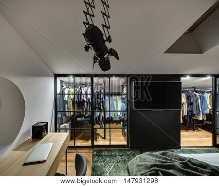 Elegant modern bedroom with cloakroom with glowing lamps, white walls. There is bed, table with laptop and chair, TV, window at the top. There are a lot of clothes in the cloakroom. Horizontal.