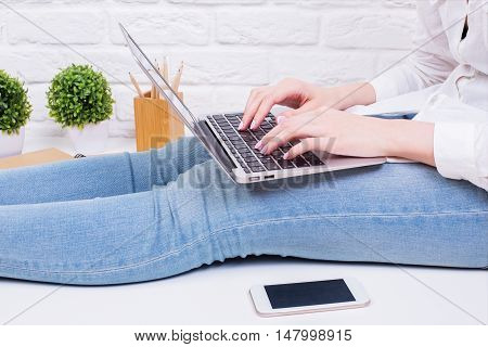 Side view of woman in jeans using laptop computer while sitting on white surface with blank smartphone decorative plants and supplies on white brick wall background. Mock up
