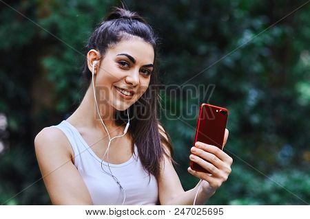 Young Girl Is Taking Picture Of Herself Using Red Cell Phone And Listening To The Music Using White 