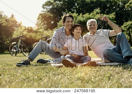 Relaxing Together. Happy Friendly United Family Sitting On The Grass Smiling And Grandpa Showing Pea