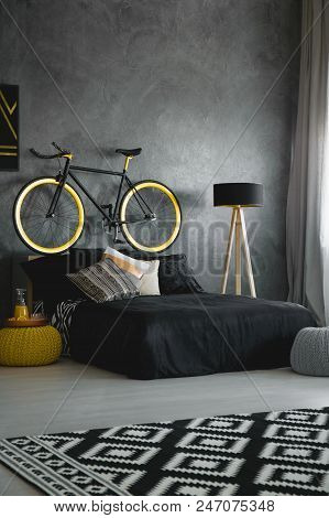 Bike Above Black Bed Against Concrete Wall In Modern Bedroom Interior With Patterned Rug. Real Photo