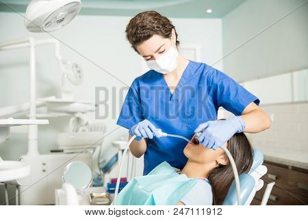 Female Dentist Using Saliva Ejector And Dental Drill While Treating Teenage Girl In Clinic
