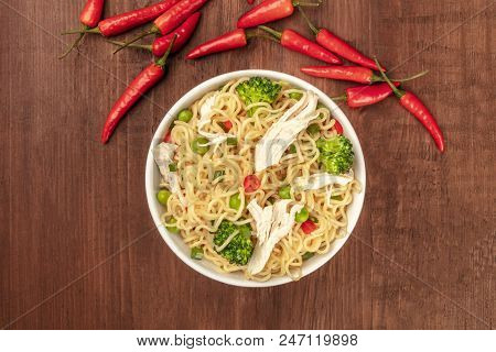 An Overhead Photo Of A Bowl Of Noodles With Chicken, Green Peas, And Broccoli, With Red Hot Chili Pe