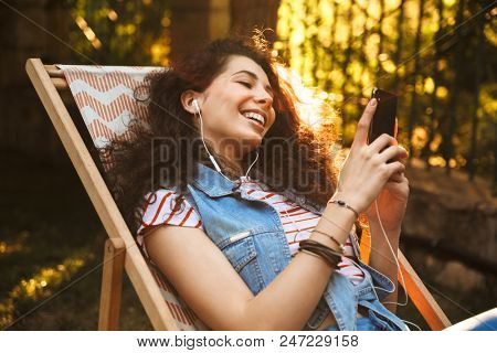Photo of happy european woman 18-20 aughing and listening to music via earphones while sitting in lounge chair during rest in park on sunny day