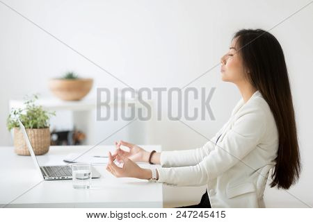 Calm Thoughtful Asian Female Employee Sitting At Office Desk Meditating, Practicing Yoga, Controllin
