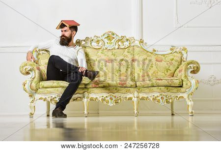 Macho Sits With Open Book On Head, Like Roof. Man With Beard And Mustache Sits On Sofa, White Wall B