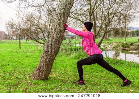 Young woman warming up and stretching the legs before running on a cold winter, autumn of fall day in an urban park. Female athlete wearing pink windbreaker, beanie, gloves and running tights