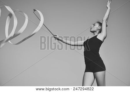 Training In Athletics. Woman Gymnast In Black Sportswear Swirl Green Ribbon On Blue Sky Background, 