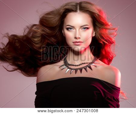 Woman high fashion model with luxurious long healthy wavy, voluminous hair in Colorful Bright Lights. Beautiful girl with chains necklace. Ledy in rock and roll style on colourful vivid red background