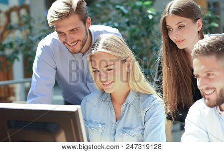 Group Of Young People In Casual Wear Sitting At The Office Desk And Discussing Something While Looki