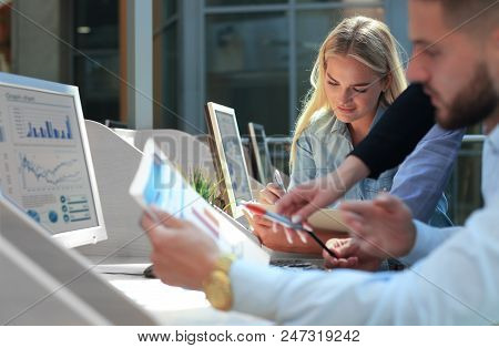 Group Of Young People In Casual Wear Sitting At The Office Desk And Discussing Something While Looki