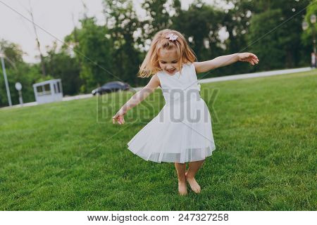 Pretty Little Cute Child Baby Girl In Light Dress Circling Around And Have Fun On Green Grass Lawn I