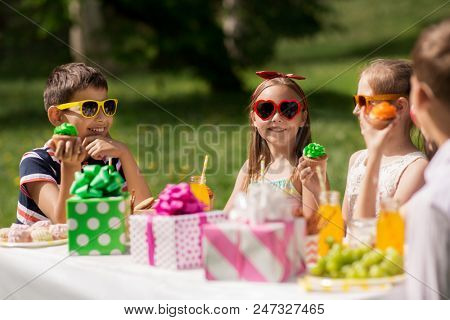 holidays, childhood and celebration concept - happy kids in sunglasses sitting at table on birthday party at summer garden and eating cupcakes