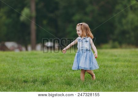Pretty Little Cute Child Baby Girl In Denim Dress Standing, Play And Have Fun On Green Grass Lawn In