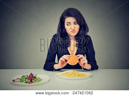 Junk Food Vs Healthy Food Choice. Young Hesitant Woman With Cheeseburger Looking At Green Vegetables