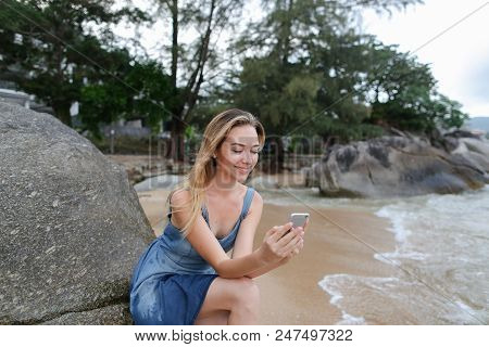 Young Girl Sitting On Sand Near Sea And Stones, Chatting By Smartphone. Concept Of Modern Technology