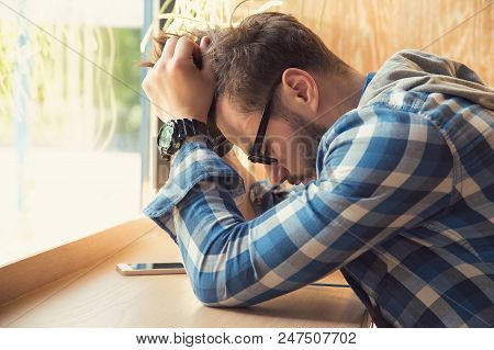 Young Depressed Man Sitting At Table With Mobile Phone In Cafe Looking Down Being Unhappy With Break