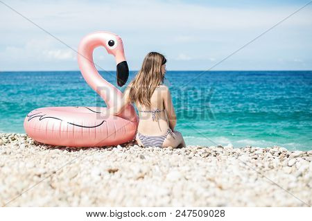 Young Woman With Bikini Sitting Close To Giant Inflated Flamingo Float On A Beach With Turquois Wate