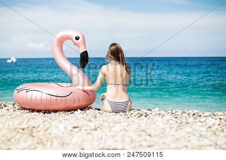 Young Woman With Bikini Sitting Close To Giant Inflated Flamingo Float On A Beach With Turquois Wate