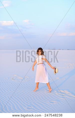 Young Caucasian Woman In White Dress Walking On Sand And Keeping Hat. Concept Of Photo Session In De