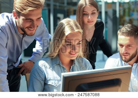 Group Of Young People In Casual Wear Sitting At The Office Desk And Discussing Something While Looki