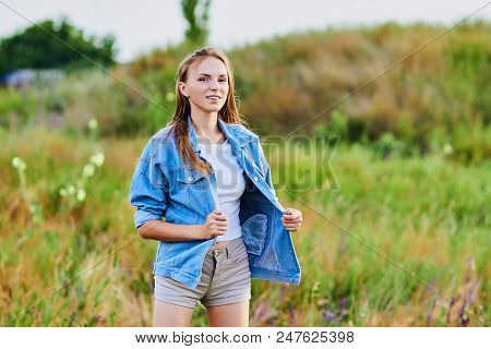 Happy Smiling Young Girl Wearing Blue Jeans Jacket And Shorts Is Standing In The Field