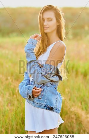 Happy Smiling Young Girl Wearing Blue Jeans Jacket And White Dress Is Standing In The Field