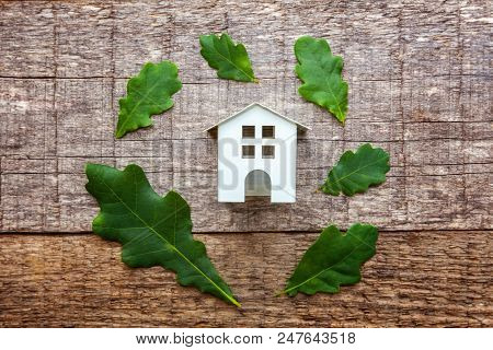 Miniature White Toy Model House With Green Oak Leaves On Wooden Backgdrop. Eco Village, Abstract Env