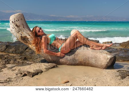 Young Sporty Suntanned Woman In Bikini And Sunglasses, Lying On Sea Drift Tree On Rocky Beach With T