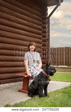 An Elderly Woman Is Sitting On A Village Bench With A Black Schnauzer Near A Wooden House In The Cou
