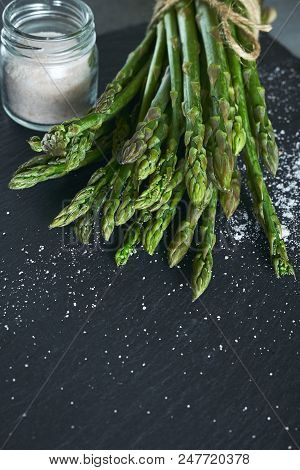 Food Background From Bunch Of Fresh Raw Garden Asparagus And Salt On Rough Black Slate Board. Ingred