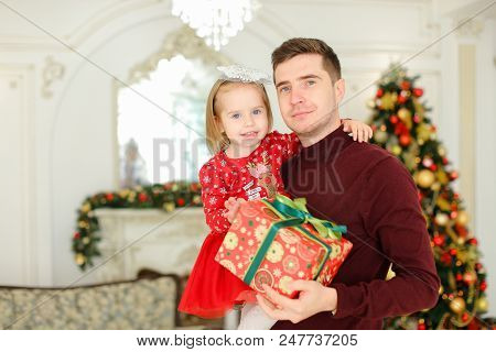 Young Happy Father Keeping Little Daughter With Present, Christmas Tree In Background. Concept Of Gi