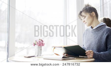 A Young Brunette Girl In A Cafe Sitting Near The Window Dressed In A Blue Pullover Is Holding A Blac