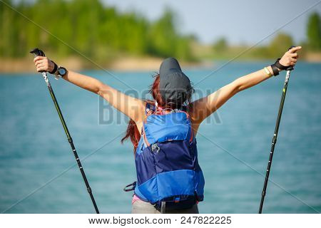 Photo of back of sporty woman with walking sticks and backpack on lake background