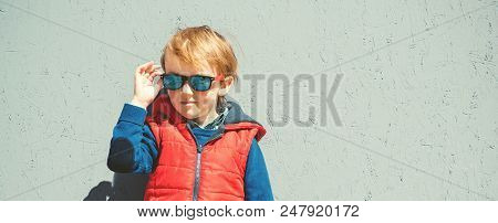 Stylish Kid In Trendy Sunglasses. Kids Fashion. Cute Little Blondy Boy In Red Jacket Standing Over G