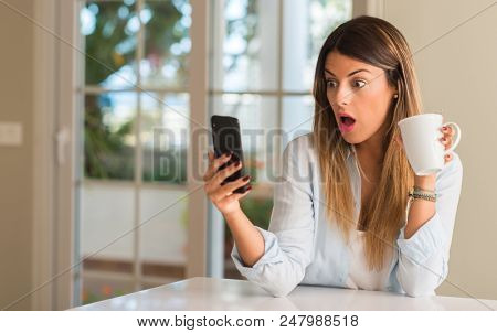 Beautiful young woman looking at the phone suprised and excited while drinking cup of coffee at home.