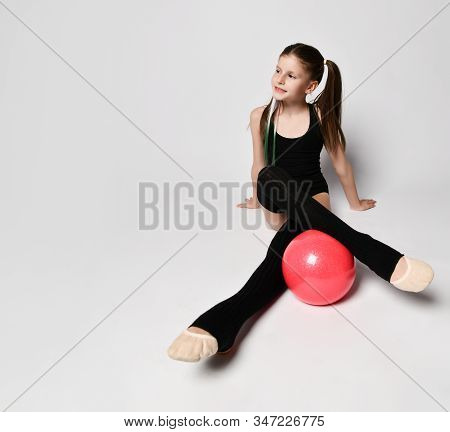 Young Smiling Girl Gymnast With Long Hair In Black Sport Body And Uppers Sitting On Floor Holding Pi