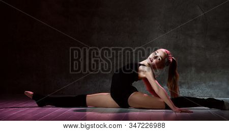 Young Smiling Girl Gymnast With Long Hair In Black Sport Body And Uppers Sitting In Twine Sideways O