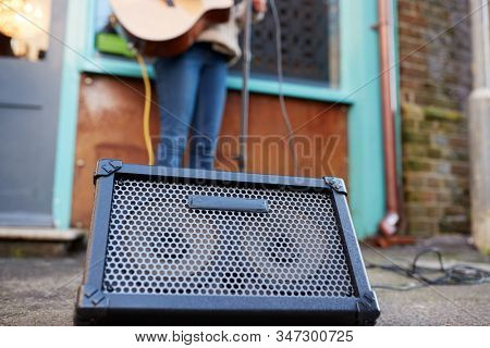 Close Up Of Amplifier As Female Musician Busks Playing Acoustic Guitar Outdoors In Street