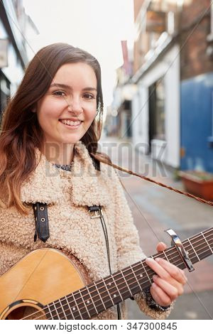 Portrait Of Young Female Musician Busking Playing Acoustic Guitar And Singing Outdoors In Street