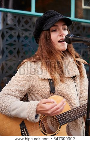 Female Musician Busking Playing Acoustic Guitar And Singing Outdoors In Street
