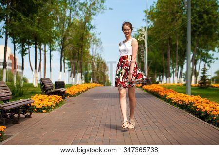 Full body portrait of a beautiful young brunette woman walking in flowers dress, summer park outdoors
