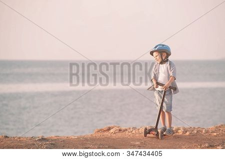 Active Handsome Kid In Sport Helmet Standing On Sea Shore With Scooter During Leisure Activity